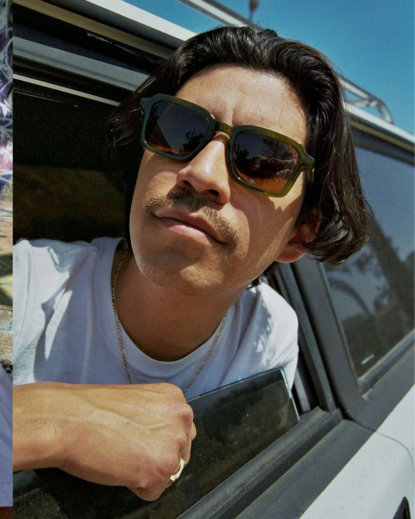 A male presenting model wearing the sunglasses and hanging out of a car window. 
